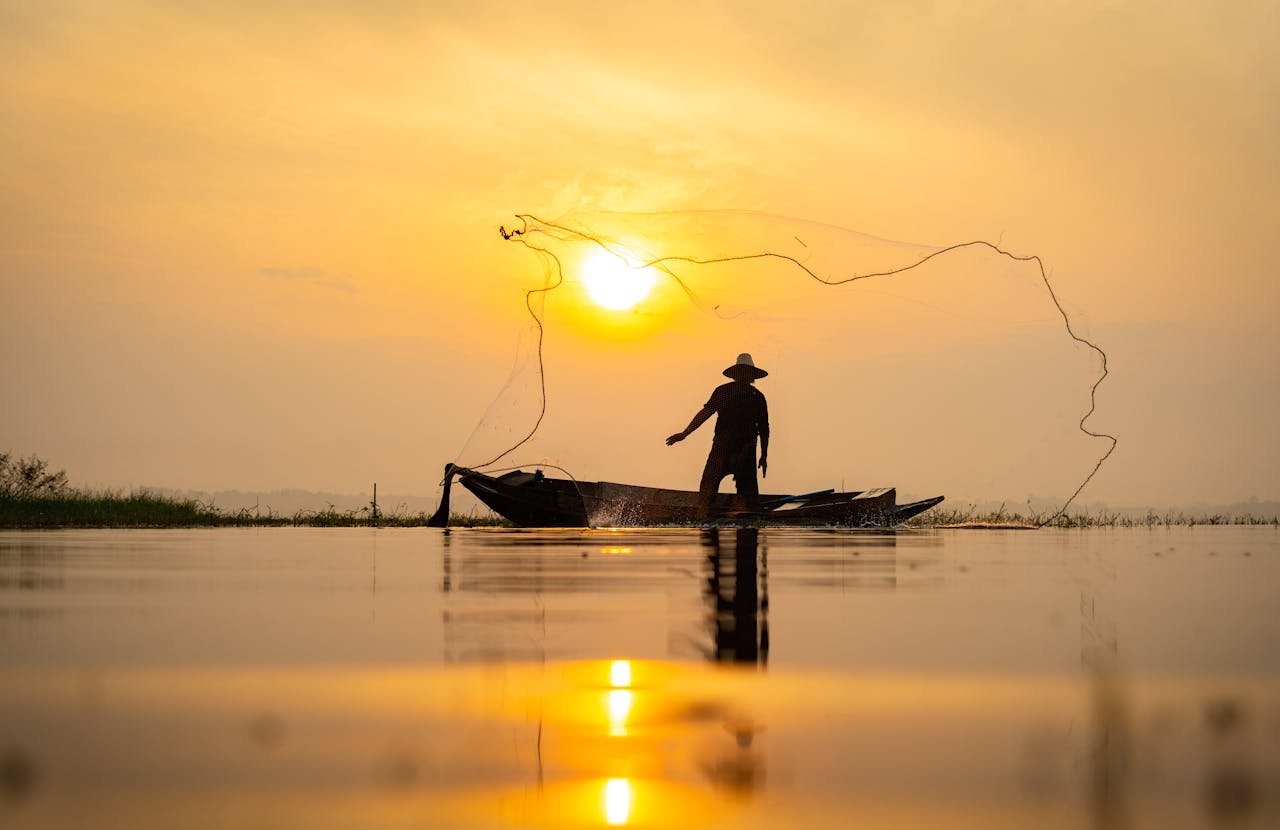 A fisherman casts his net at sunrise in Bangkok, Thailand, creating a silhouette on tranquil waters.